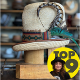 Luna Straw Top Hat with a woven natural straw finish, decorated with a colorful patterned band and vibrant feathers, displayed on a wooden stand in a hat shop.