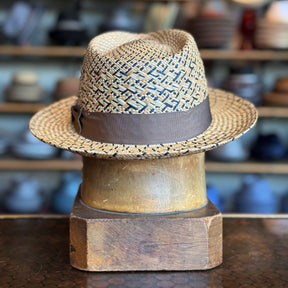 Back of Maize Panama Fedora hat with braided straw pattern and dark brown leather band, displayed on a wooden stand against a blurred background of hats and accessories.