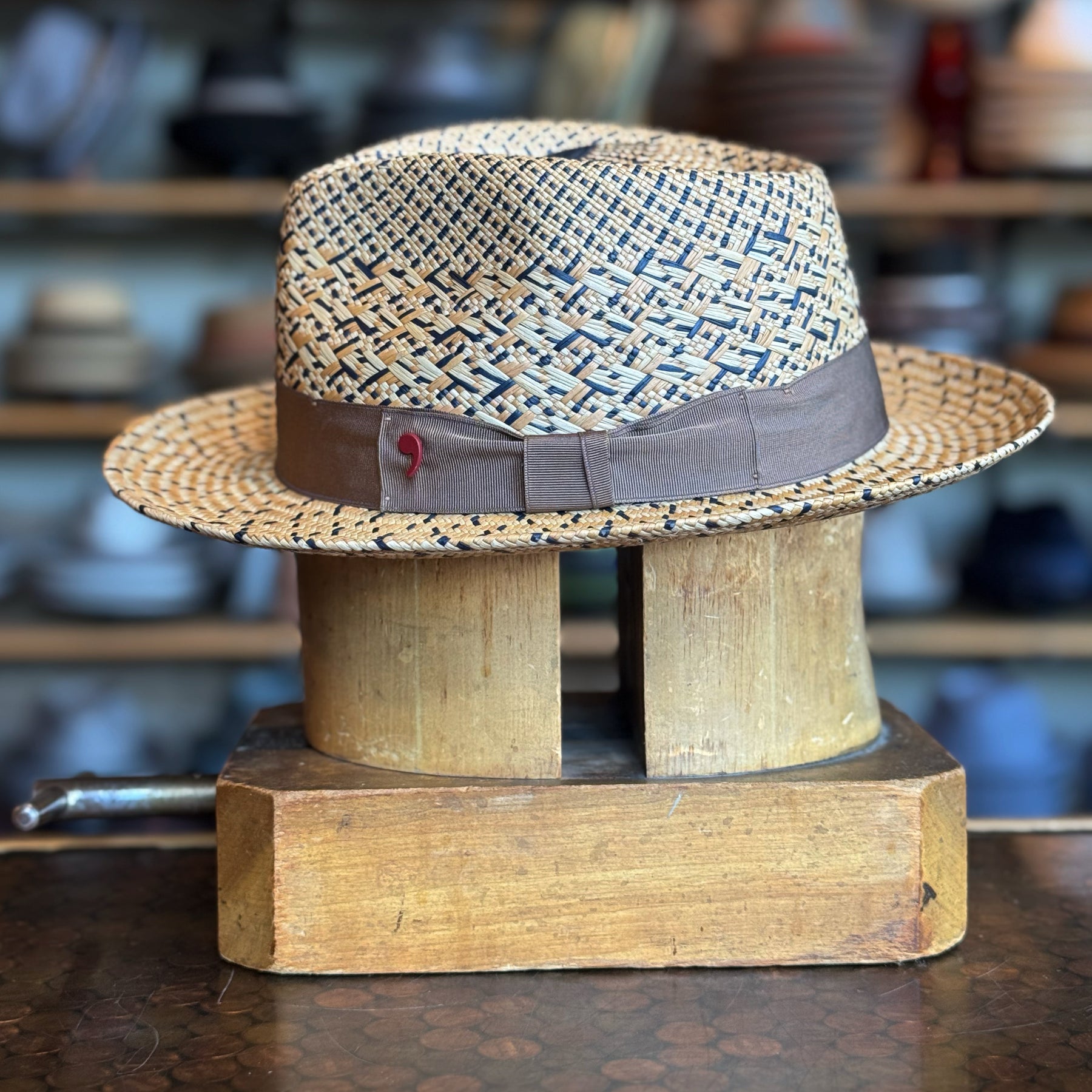 Maize Panama Fedora with woven straw pattern and dark brown fabric band, displayed on a wooden hat block in a shop setting with blurred shelves in the background.