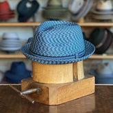 Mannes hat in a woven blue pattern with a black rope trim, displayed on a wooden hat block in a shop with various hats in the background.