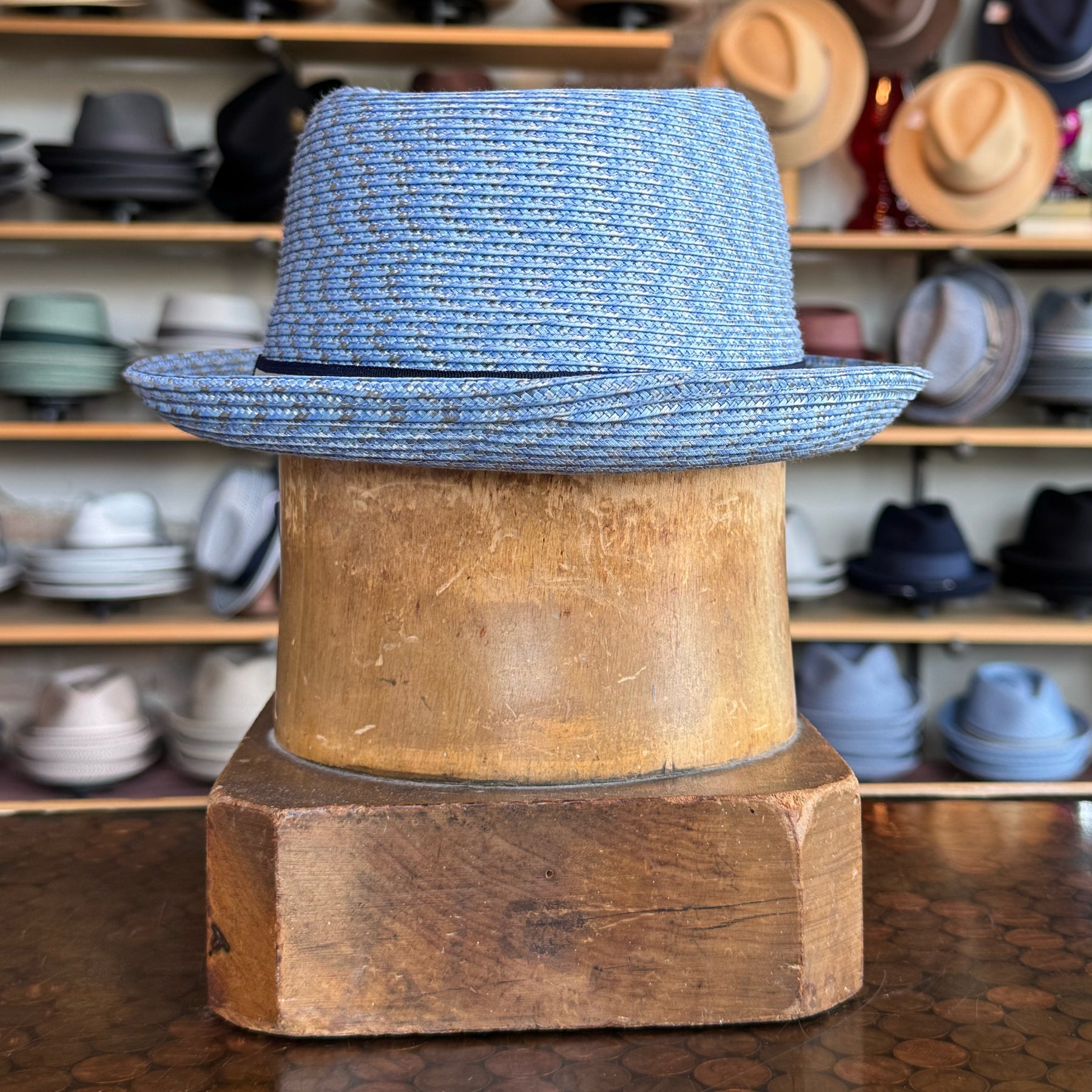 Mannes light blue woven hat with a subtle brim and black trim displayed on a wooden hat stand in front of a shelf filled with various hats.