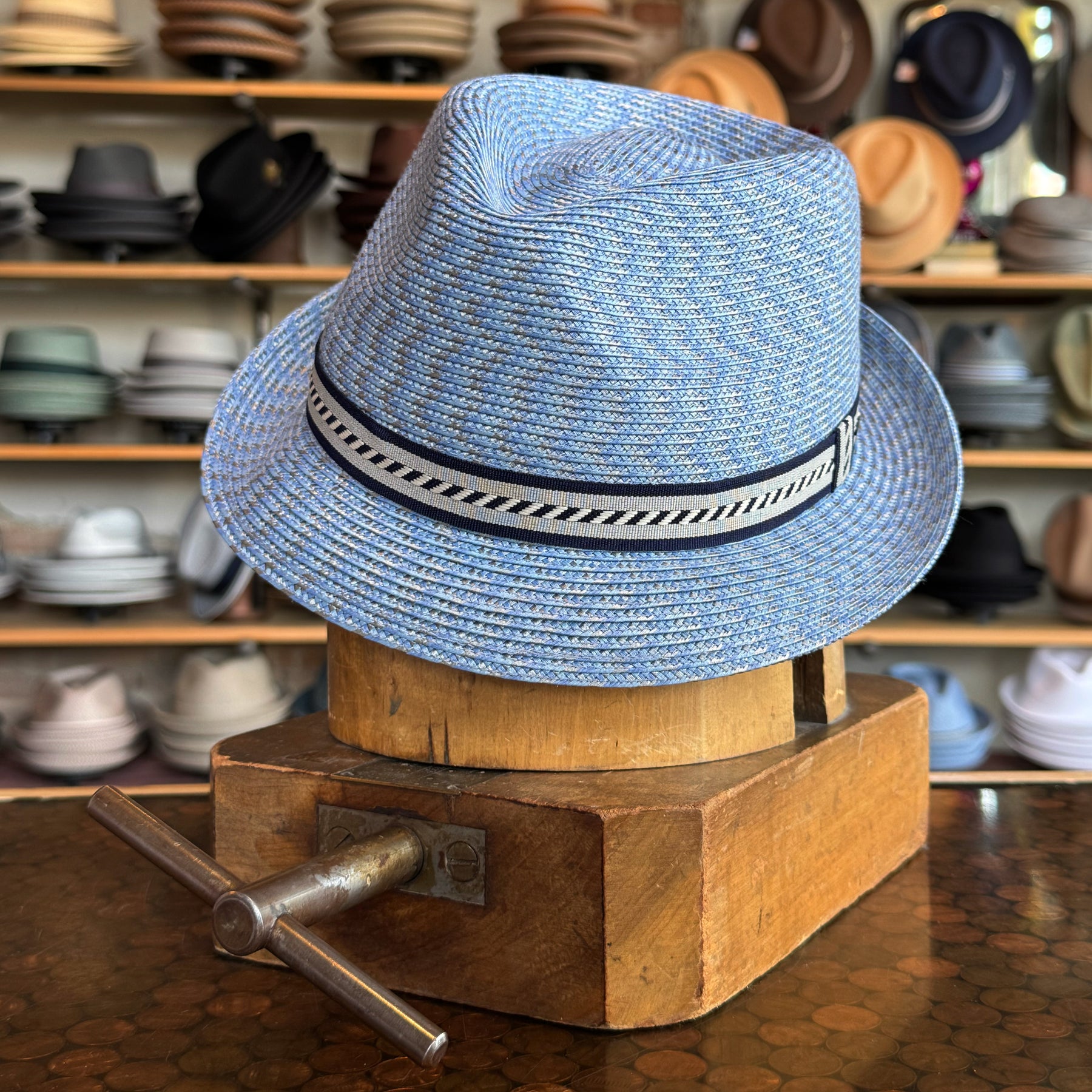 Mannes blue woven fedora hat with a black and white patterned band displayed on a wooden hat block in a hat shop with various hats in the background.