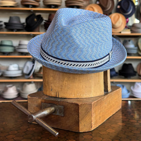 A blue woven Mannes hat with a patterned black and white band displayed on a vintage wooden hat block with shelves of various hats blurred in the background.
