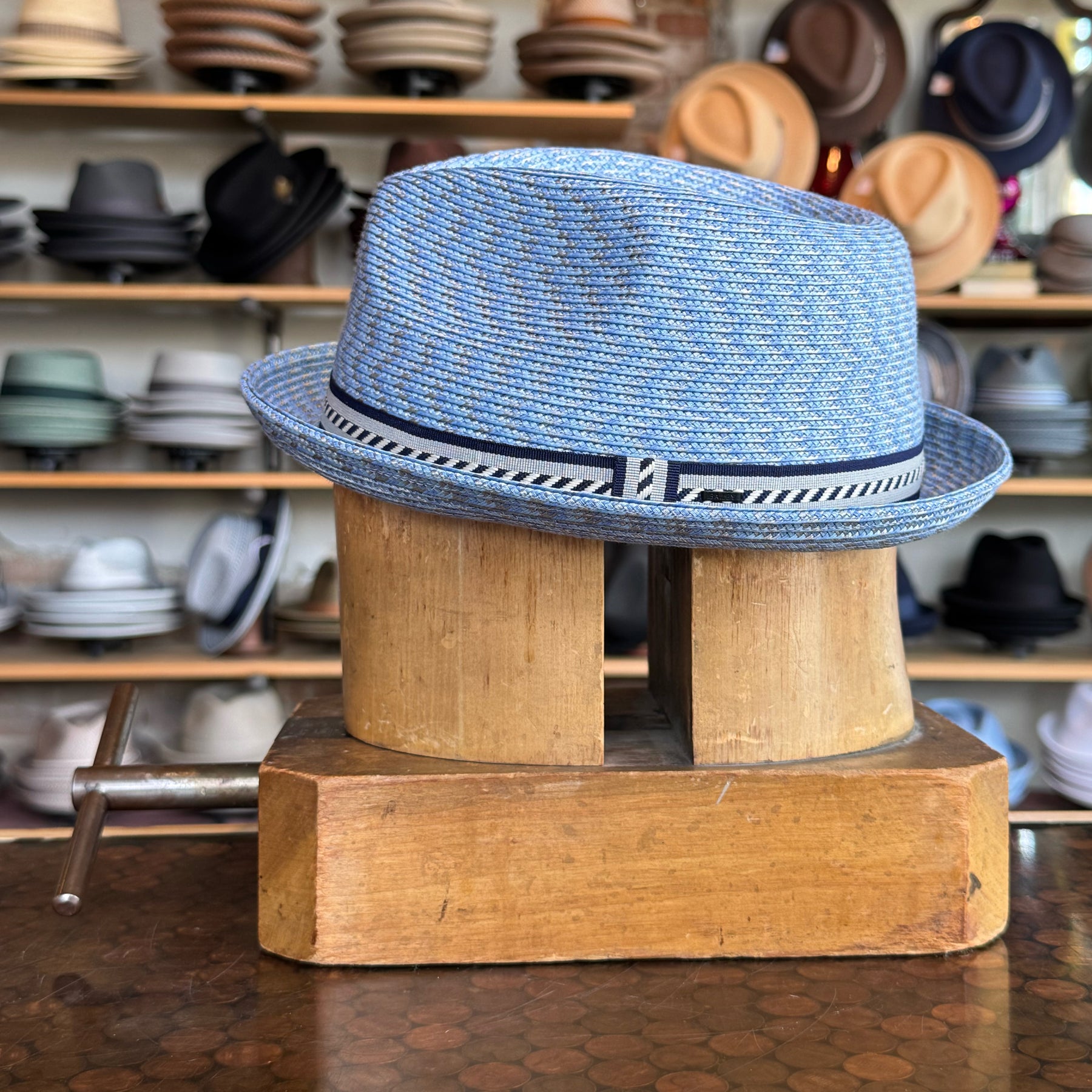 Mannes blue woven fedora with a patterned band around the crown, displayed on a wooden hat block with various hats in the background.