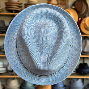 Mannes straw fedora hat in a light blue woven pattern, displayed in front of a shelf filled with various hats, showcasing a classic brim and teardrop crown design.