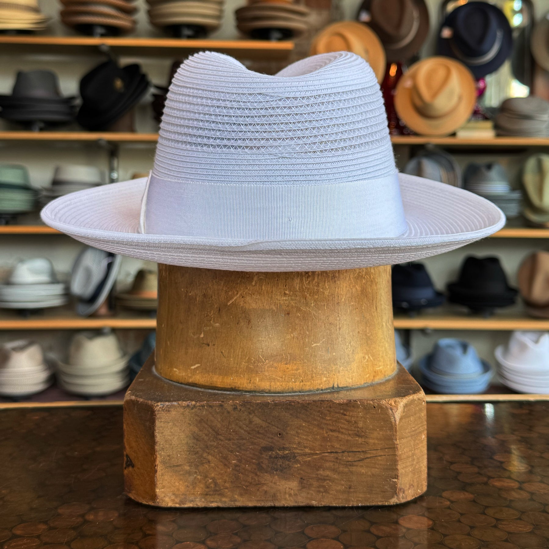 Max white fedora hat with a wide brim and ribbon band displayed on a wooden stand in a hat shop showcasing various styles in the background