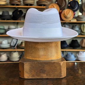 Max white fedora hat with a wide brim and ribbon band displayed on a wooden stand in a hat shop showcasing various styles in the background