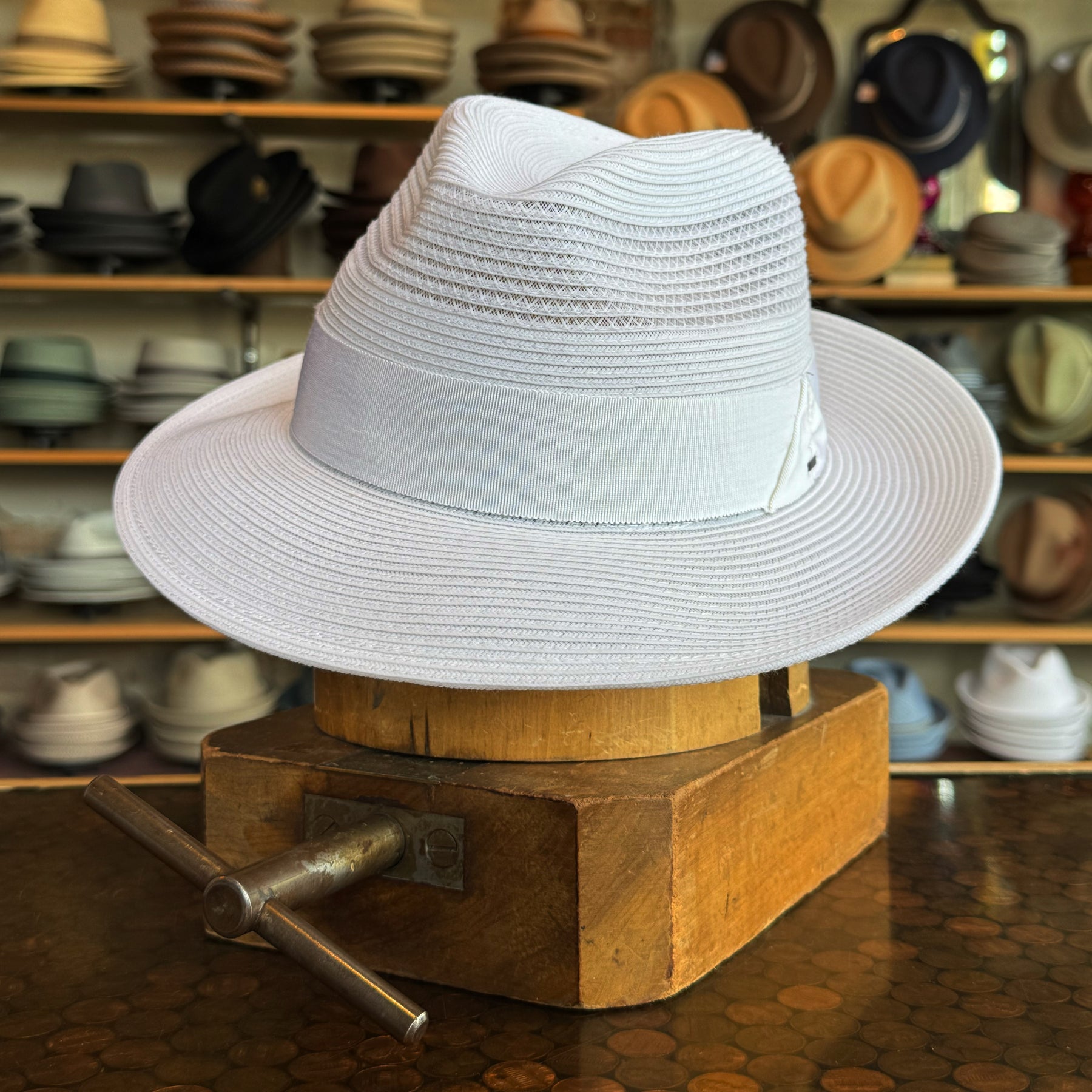 White fedora-style hat named Max with a textured woven design and a wide brim, displayed on a wooden hat stretcher in a shop with various hats in the background