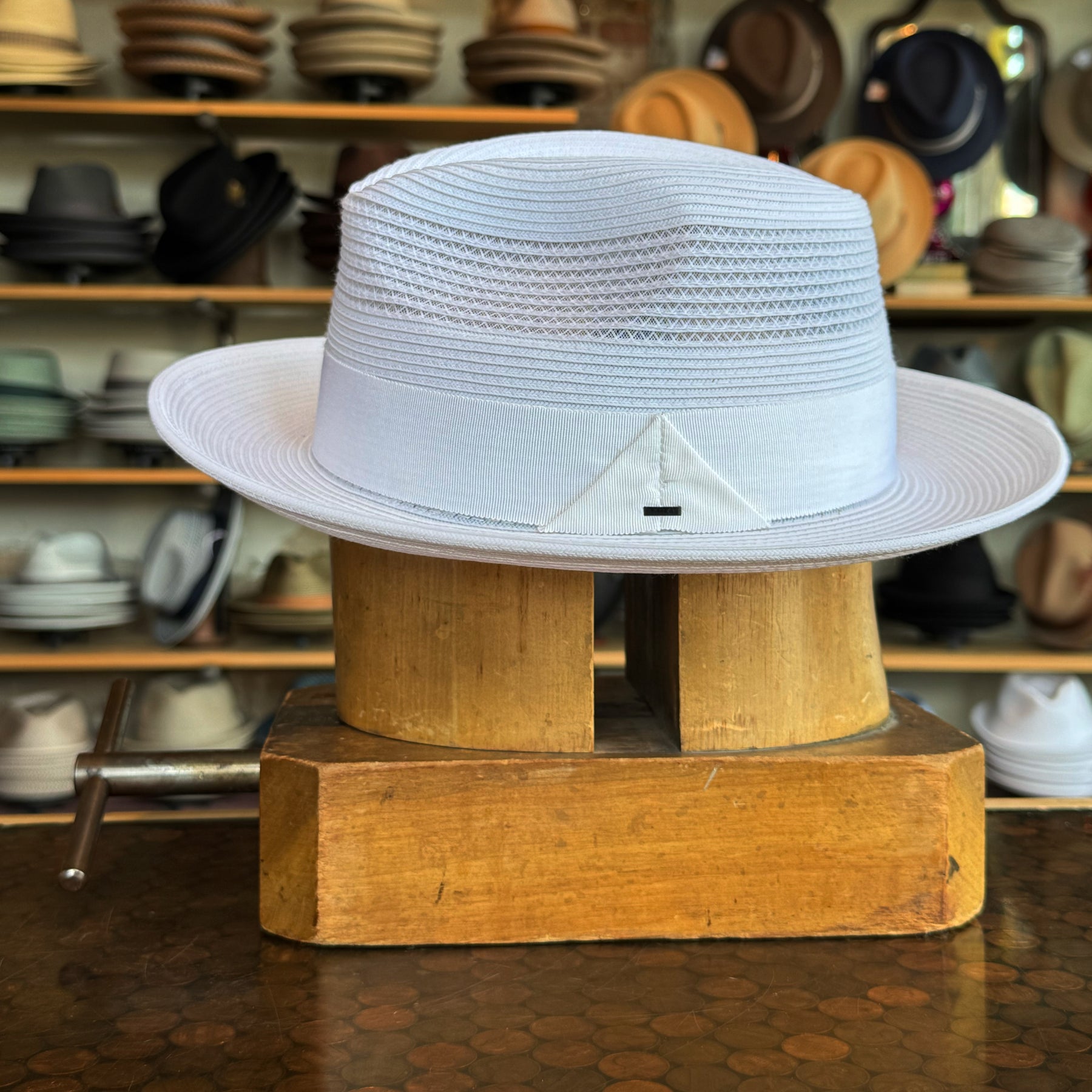 White Max fedora-style hat with woven texture and wide brim displayed on a wooden hat block in a shop with various hats in the background