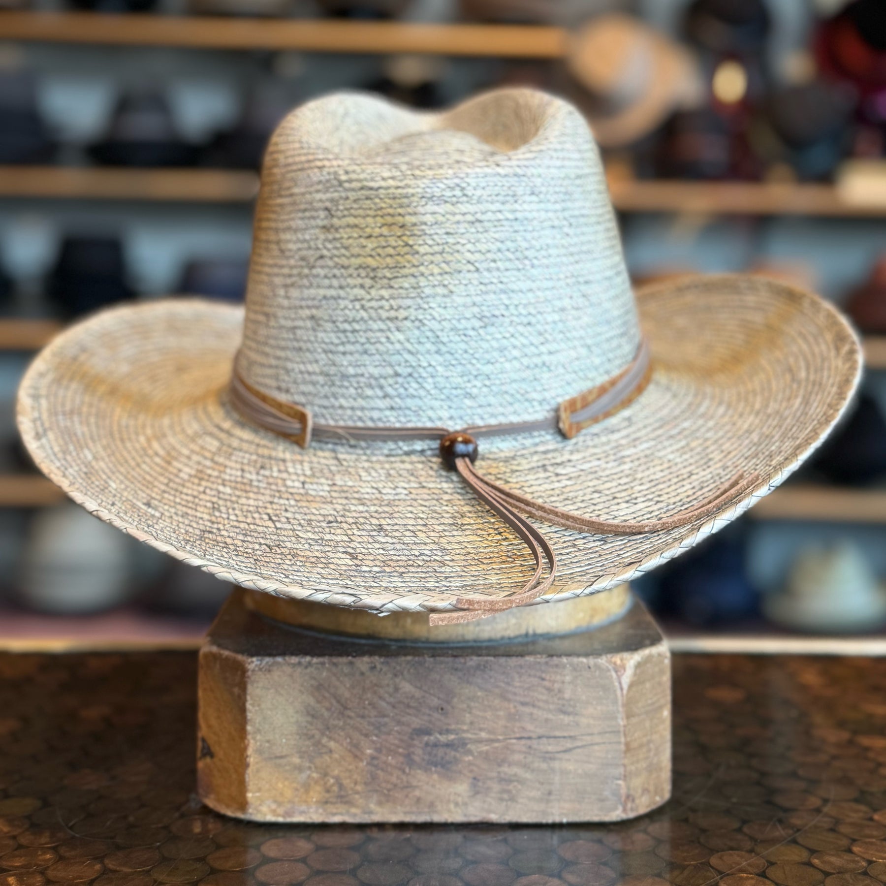 Monterrey Bay straw hat with a wide brim and leather band detail, displayed on a wooden stand in a rustic hat shop setting.