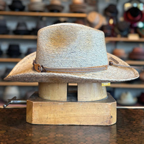 Monterrey Bay wide-brim straw hat with a leather band accent, displayed on a wooden hat block in a shop filled with various hats in the background.