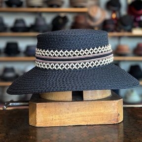 Noreen black woven wide-brim hat featuring intricate cream-colored decorative bands, displayed on a wooden hat block with blurred hats in the background.
