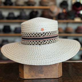 Noreen natural woven straw wide-brim hat with black and beige patterned band, featuring a small round logo accent, displayed on a wooden stand in a hat shop.