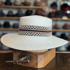 Noreen natural straw wide-brim hat with decorative patterned band, displayed on a wooden stand in a hat shop setting.