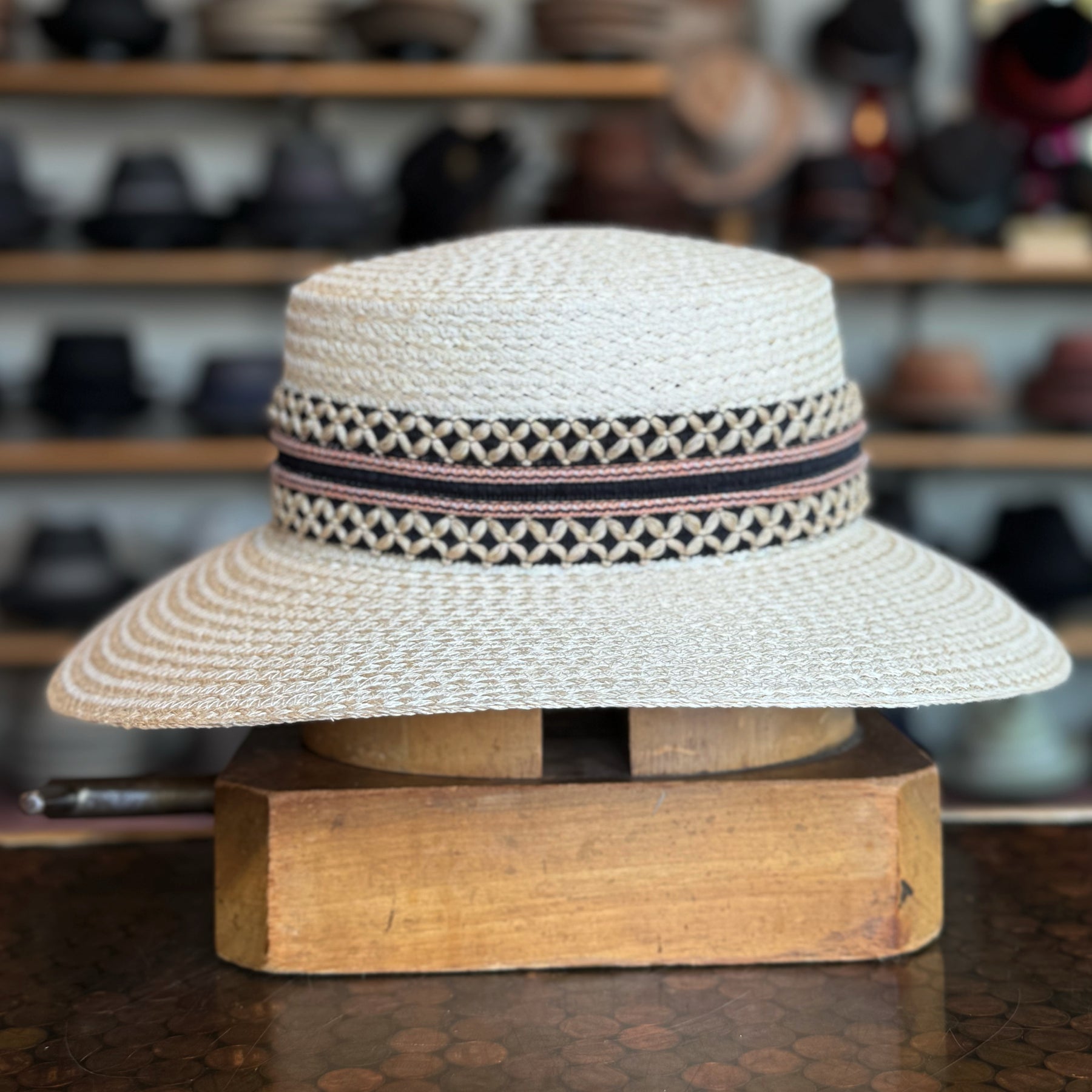 Noreen natural woven straw hat with wide brim and decorative black and pink patterned band displayed on a wooden stand in a hat shop.