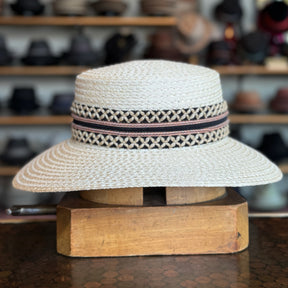 Noreen natural woven straw hat with wide brim and decorative black and pink patterned band displayed on a wooden stand in a hat shop.
