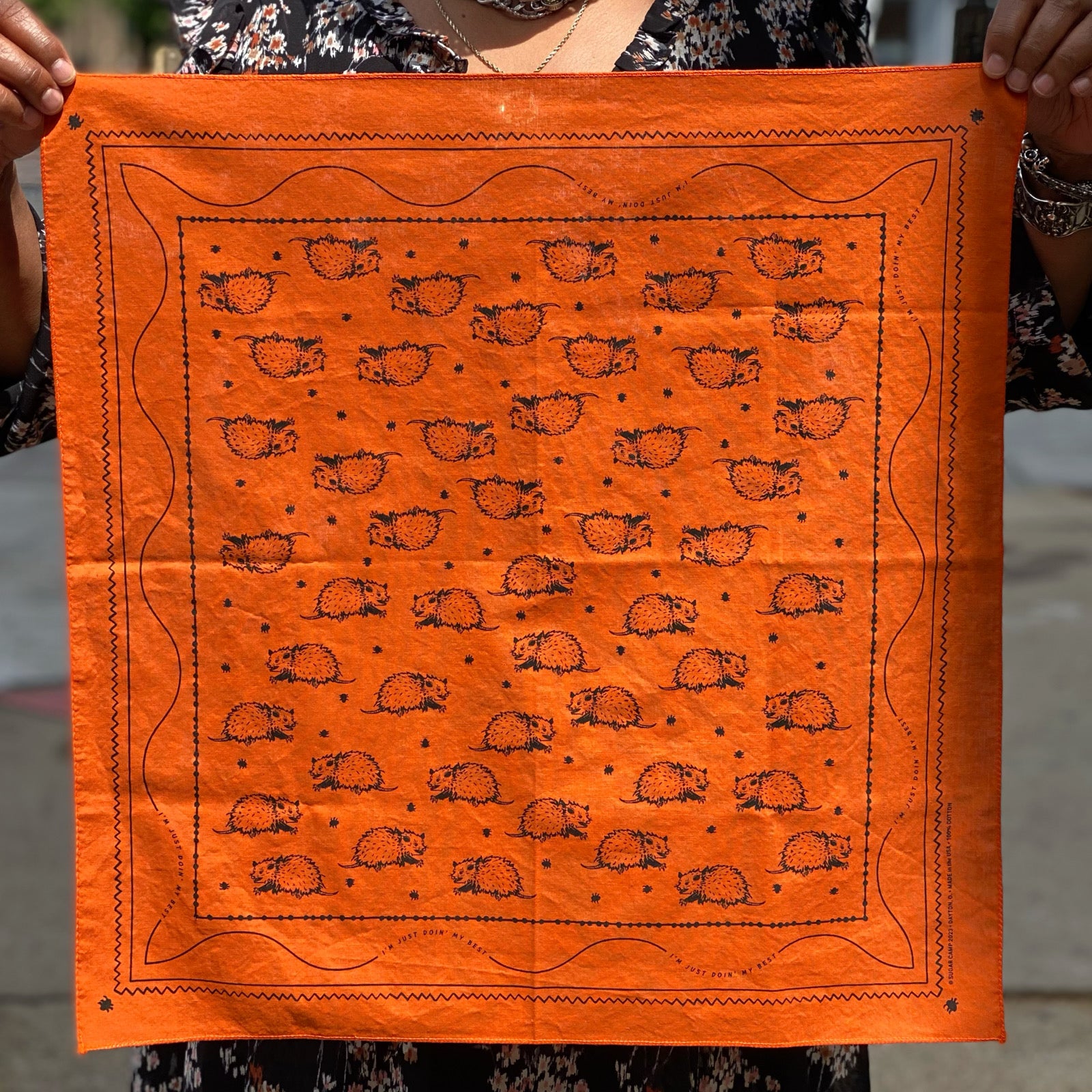 Opossum Bandana in burnt orange featuring a repeating black opossum pattern with decorative border, held by a person outdoors.