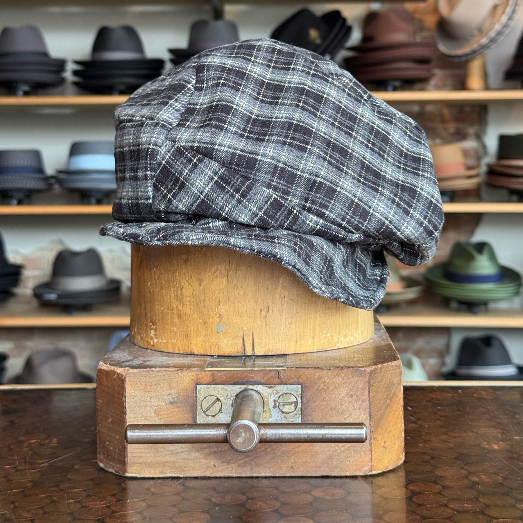 Plaid Funk Master Cap in a black and gray plaid pattern displayed on a wooden hat block with various other hats blurred in the background.