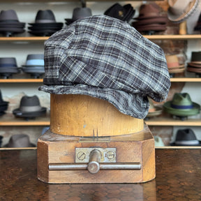 Plaid Funk Master Cap in a black and gray plaid pattern displayed on a wooden hat block with various other hats blurred in the background.