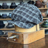 Plaid Funk Master Cap displayed on a wooden hat block, featuring a black and gray plaid pattern in a classic newsboy cap style, with hats blurred in the background.