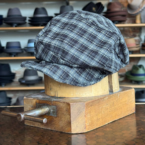 Plaid Funk Master Cap displayed on a wooden hat block, featuring a black and gray plaid pattern in a classic newsboy cap style, with hats blurred in the background.