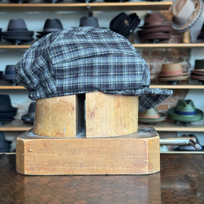 Plaid Funk Master Cap in muted dark colors displayed on a vintage wooden hat block with shelves of various other hats blurred in the background.