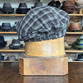 Plaid Funk Master Cap in a black and gray plaid pattern displayed on a wooden hat block with various other hats blurred in the background.