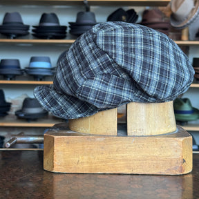 Plaid Funk Master Cap in dark plaid pattern displayed on wooden hat block, showcasing a classic newsboy style hat among various brim hats in the background.