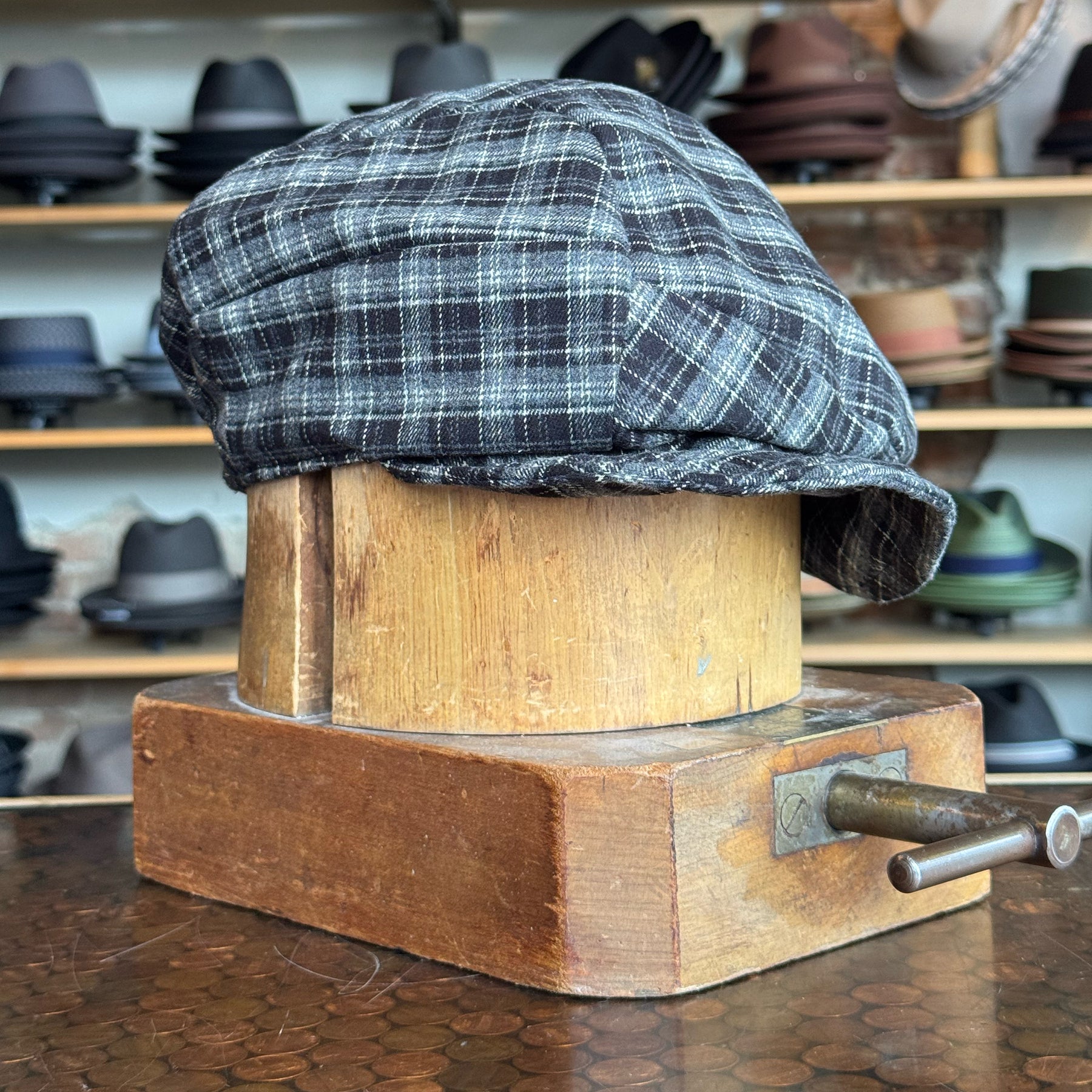 Plaid Funk Master Cap displayed on a wooden hat block, featuring a black and gray plaid pattern, surrounded by various hats in the background.