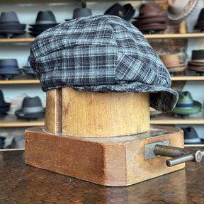 Plaid Funk Master Cap displayed on a wooden hat block, featuring a black and gray plaid pattern, surrounded by various hats in the background.