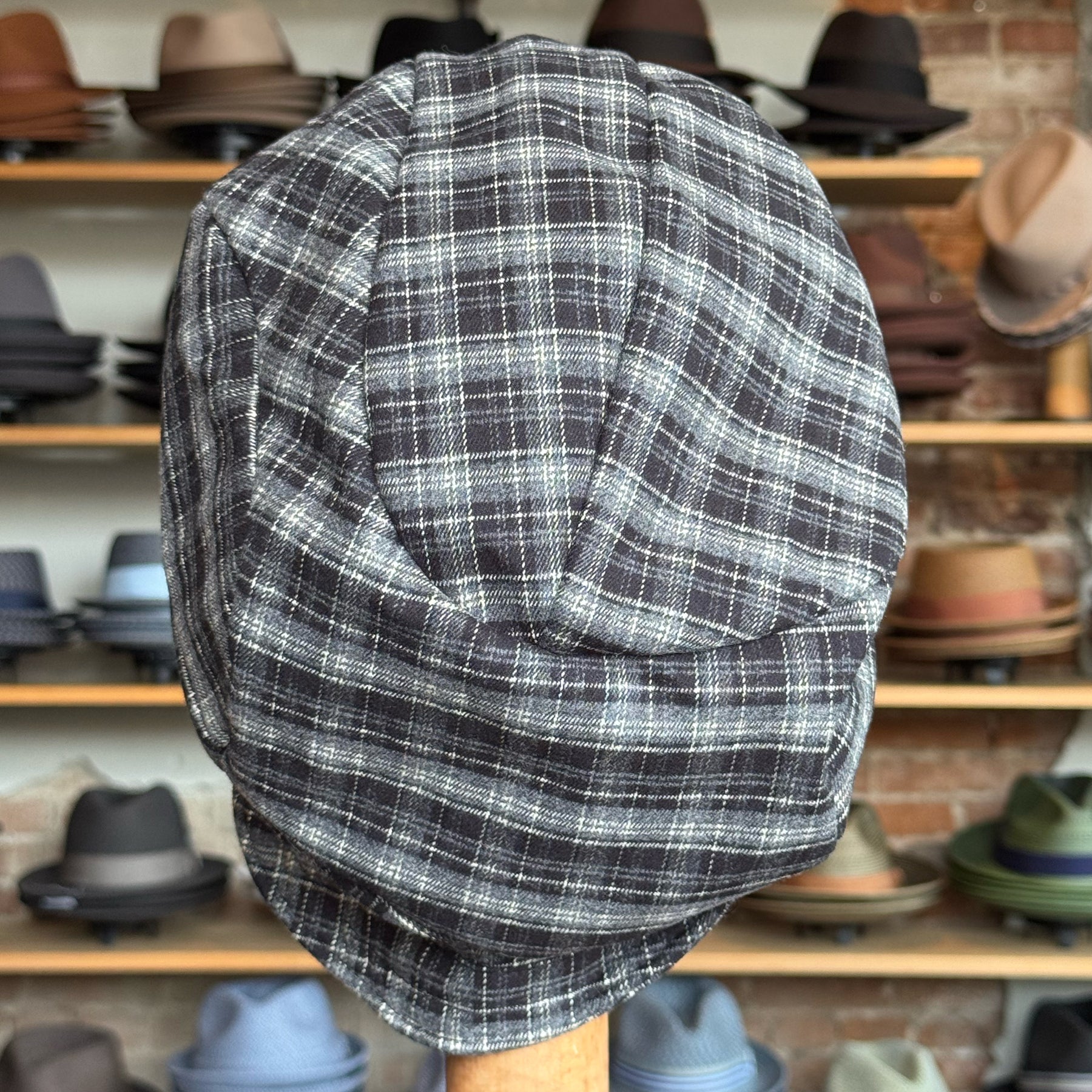 Plaid Funk Master Cap in black and grey plaid pattern displayed on a stand with various other hats blurred in the background, showcasing a stylish newsboy cap design.