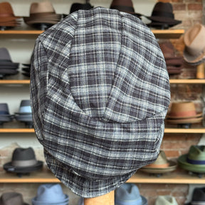 Plaid Funk Master Cap in black and grey plaid pattern displayed on a stand with various other hats blurred in the background, showcasing a stylish newsboy cap design.