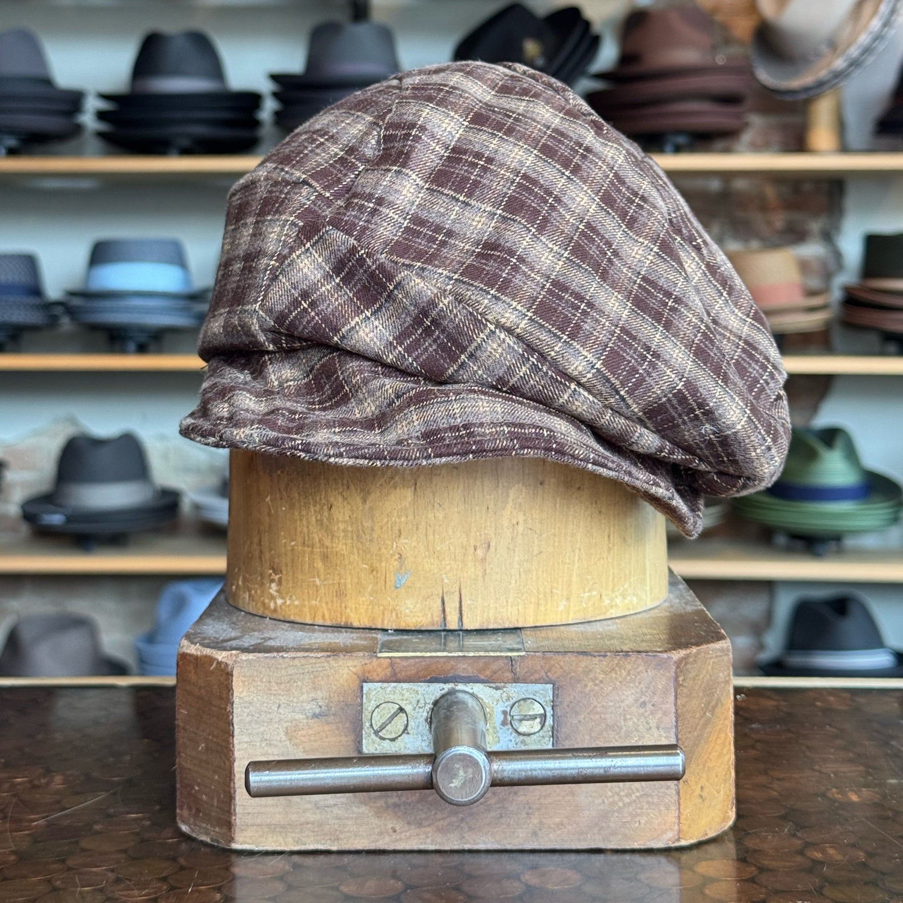 Brown plaid newsboy style flat cap displayed on a wooden hat block with various hats in the background, showcasing the Plaid Funk Master Cap.