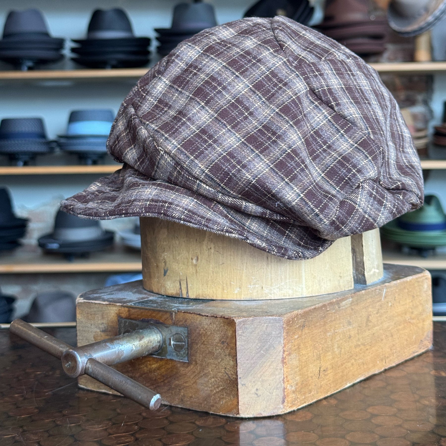 Plaid Funk Master Cap in brown plaid fabric displayed on a wooden hat block, surrounded by various hats on shelves in the background.