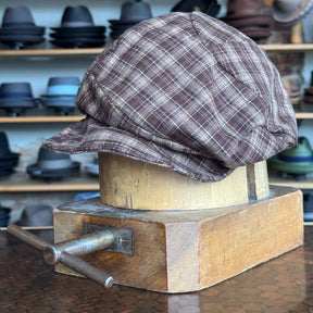Plaid Funk Master Cap in brown plaid fabric displayed on a wooden hat block, surrounded by various hats on shelves in the background.