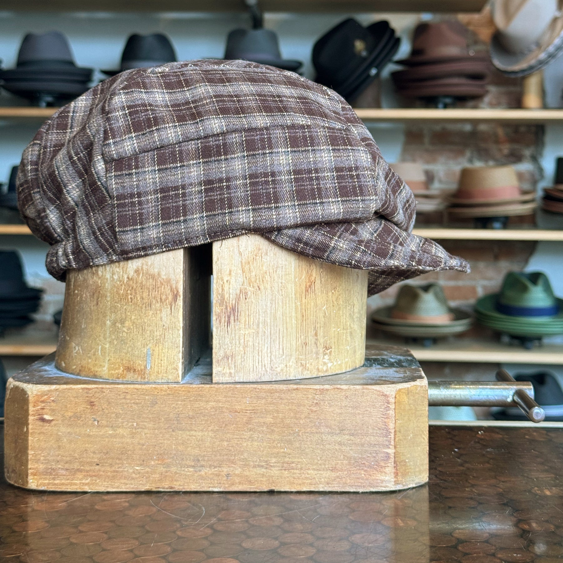 Plaid Funk Master Cap displayed on a wooden hat stand with a brown plaid pattern, showcased in a rustic hat shop featuring various styles on shelves in the background.