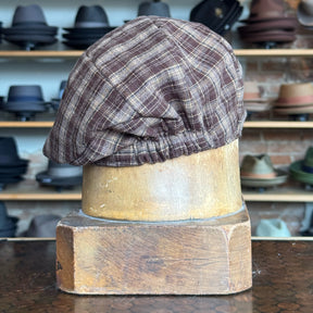 Plaid Funk Master Cap in brown plaid fabric displayed on a wooden hat block with shelves of various hats blurred in the background, showcasing a classic newsboy style.
