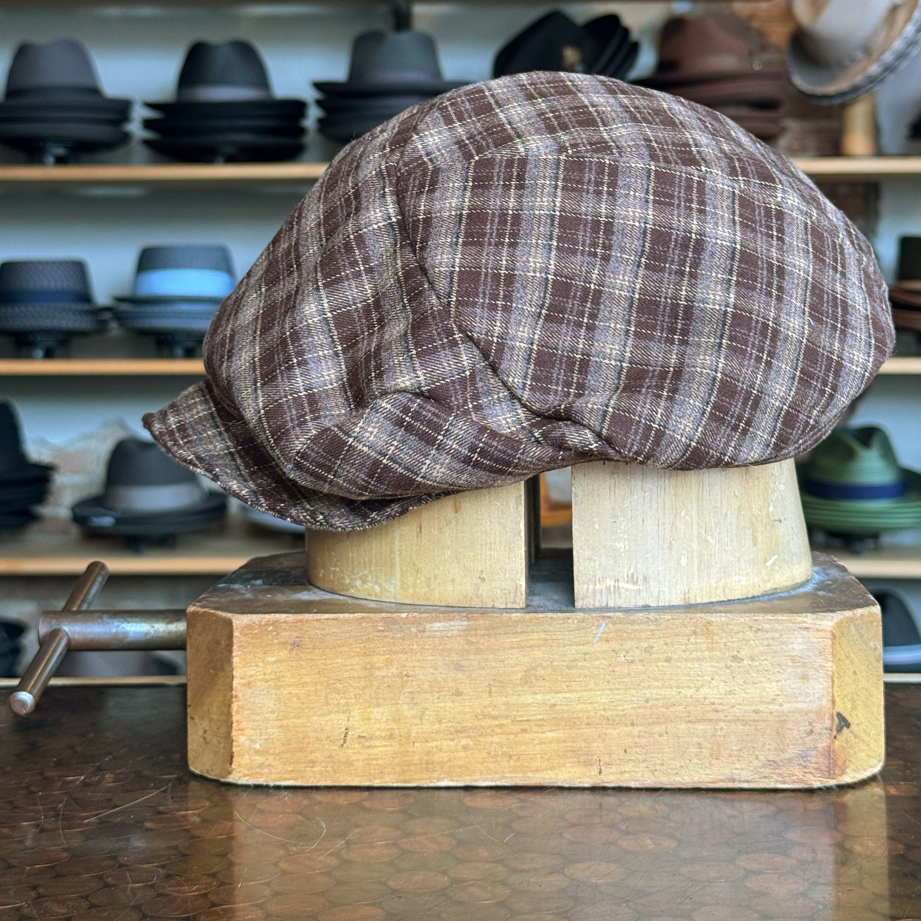 Plaid Funk Master Cap in brown plaid fabric displayed on a wooden hat block, set against a background of various hats on shelves in a hat shop.