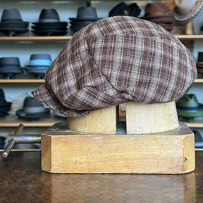 Plaid Funk Master Cap in brown plaid fabric displayed on a wooden hat block, set against a background of various hats on shelves in a hat shop.