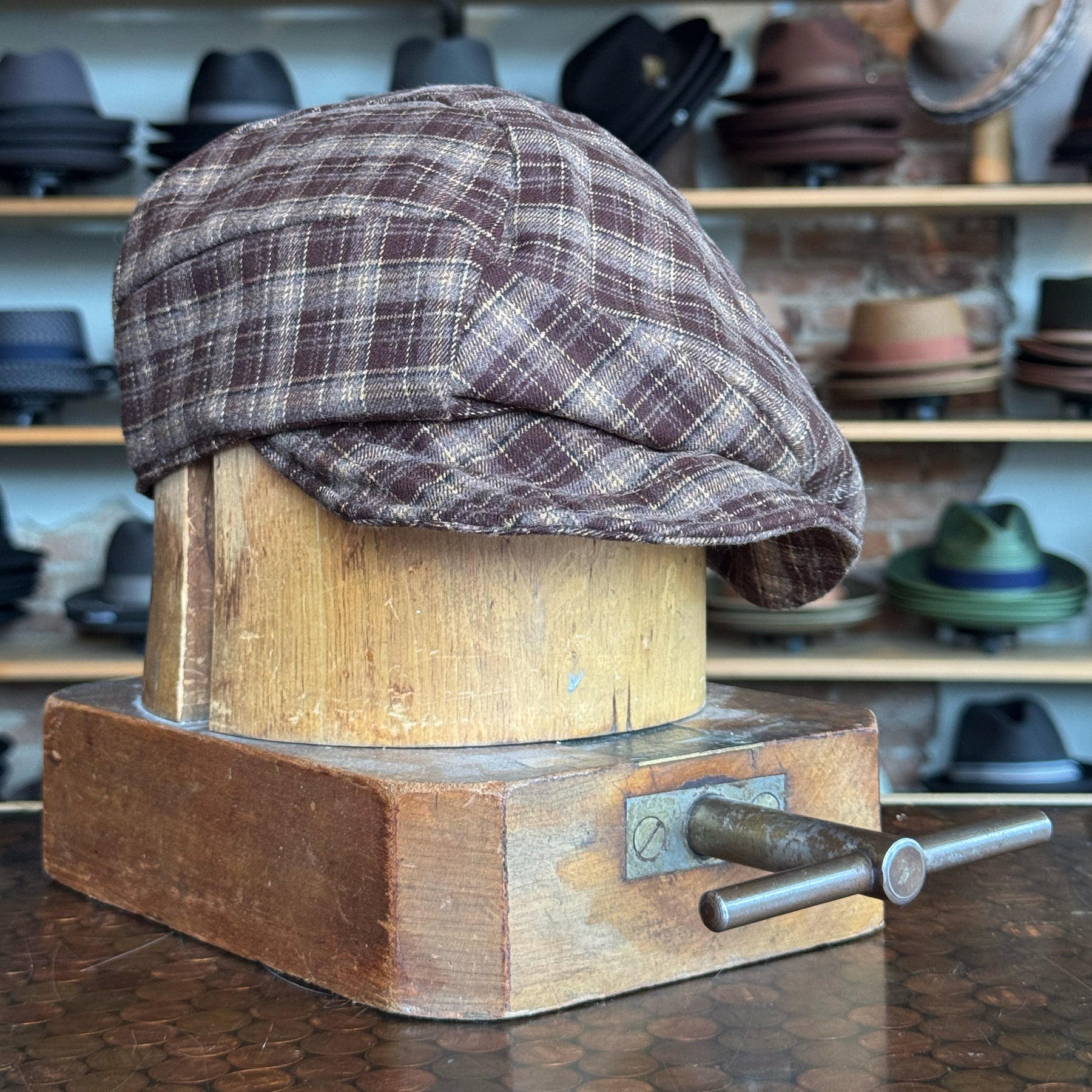 Plaid Funk Master Cap in brown plaid fabric displayed on a wooden hat block with shelves of various hats in the background, showcasing a vintage newsboy style cap.