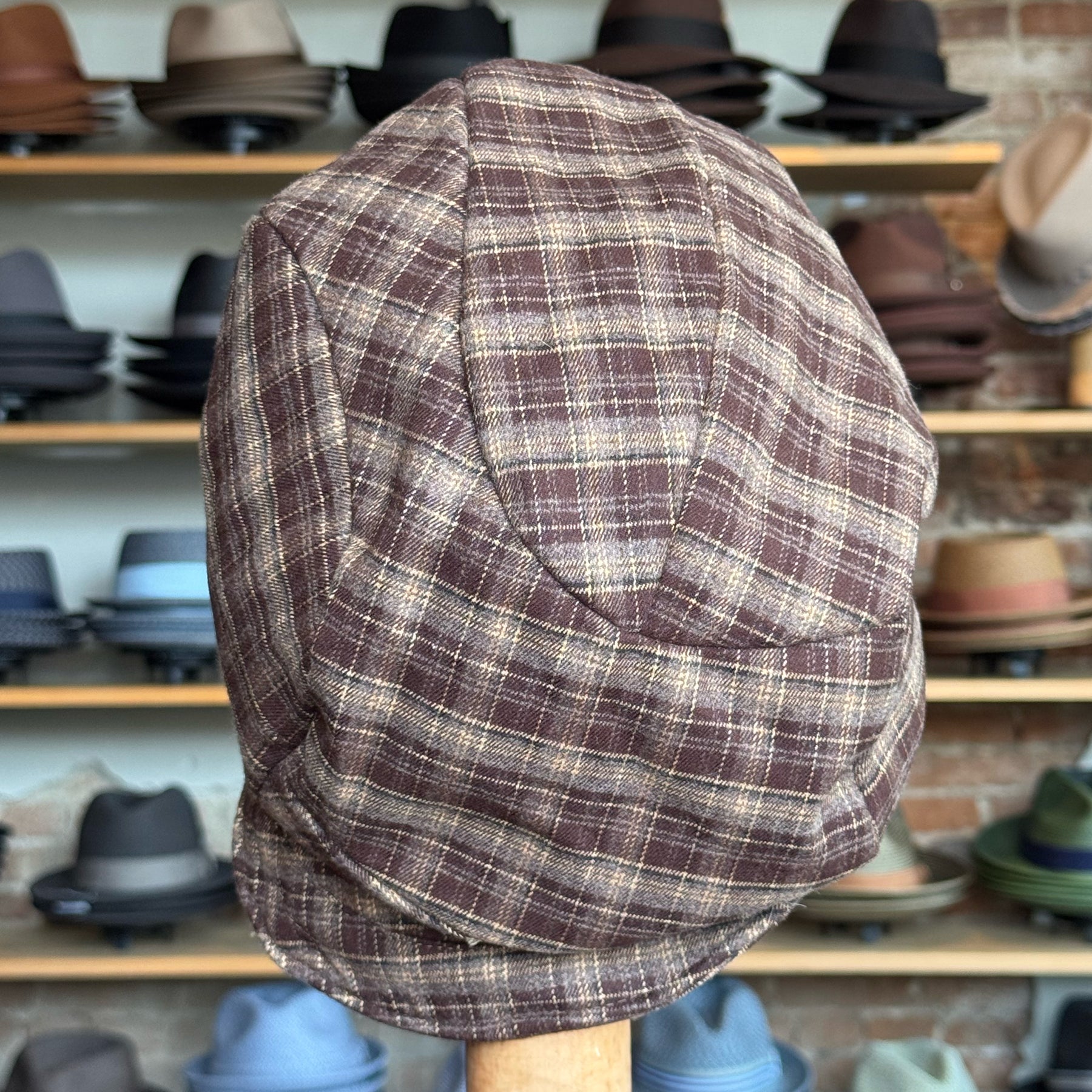 Plaid Funk Master Cap in a brown and beige checkered pattern displayed on a wooden stand with various hats in the background.