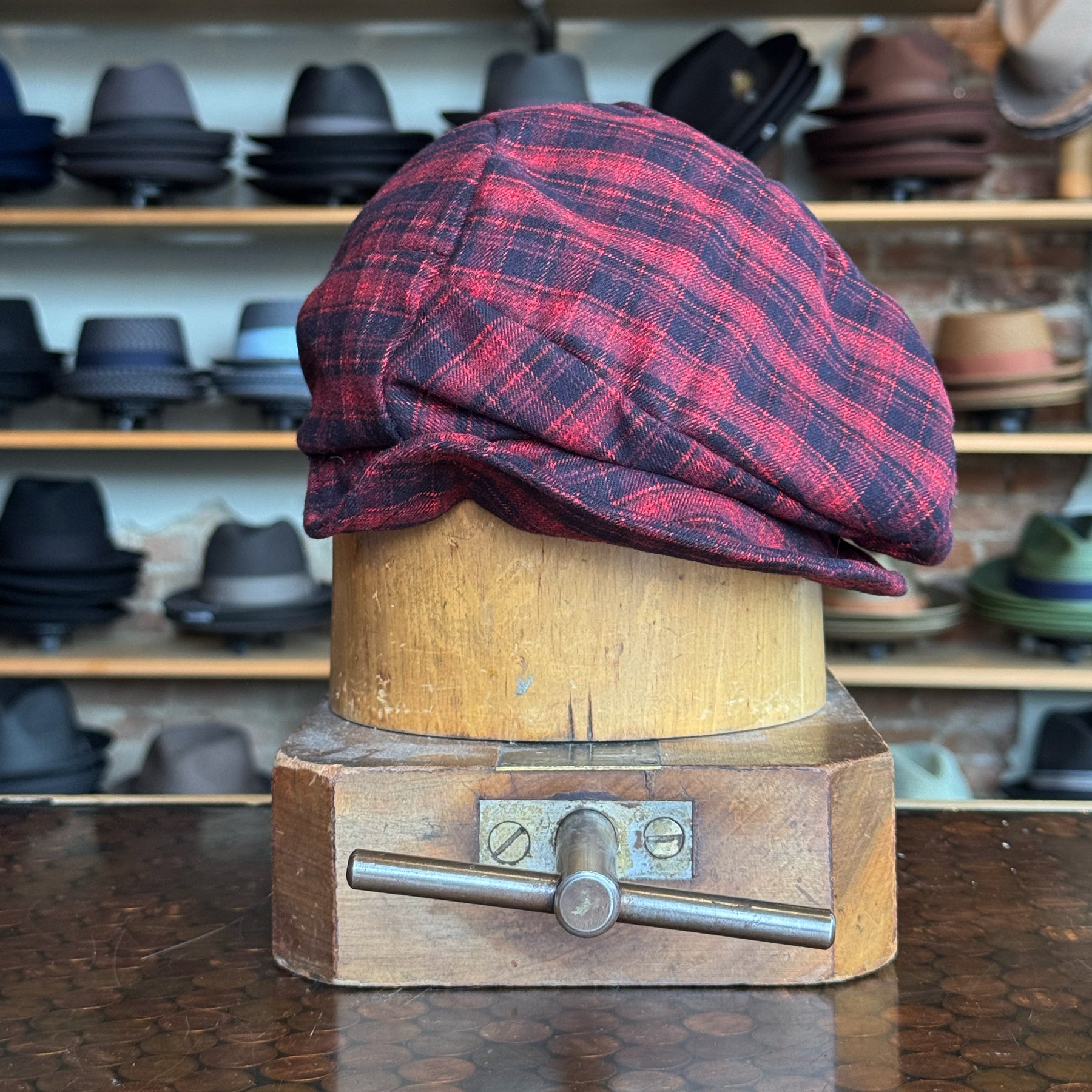Plaid Funk Master Cap in a red and black plaid pattern displayed on a vintage wooden hat block, surrounded by various styles of hats on shelves in the background.