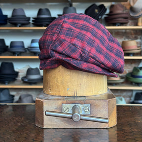 Plaid Funk Master Cap in a red and black plaid pattern displayed on a vintage wooden hat block, surrounded by various styles of hats on shelves in the background.