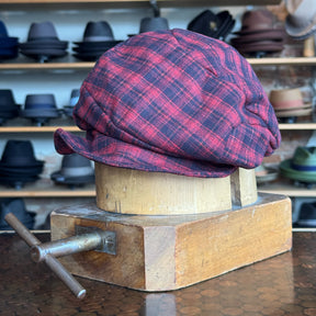 Plaid Funk Master Cap in red and black plaid pattern displayed on a wooden hat block with shelves of various hats in the background, showcasing a classic newsboy style cap.