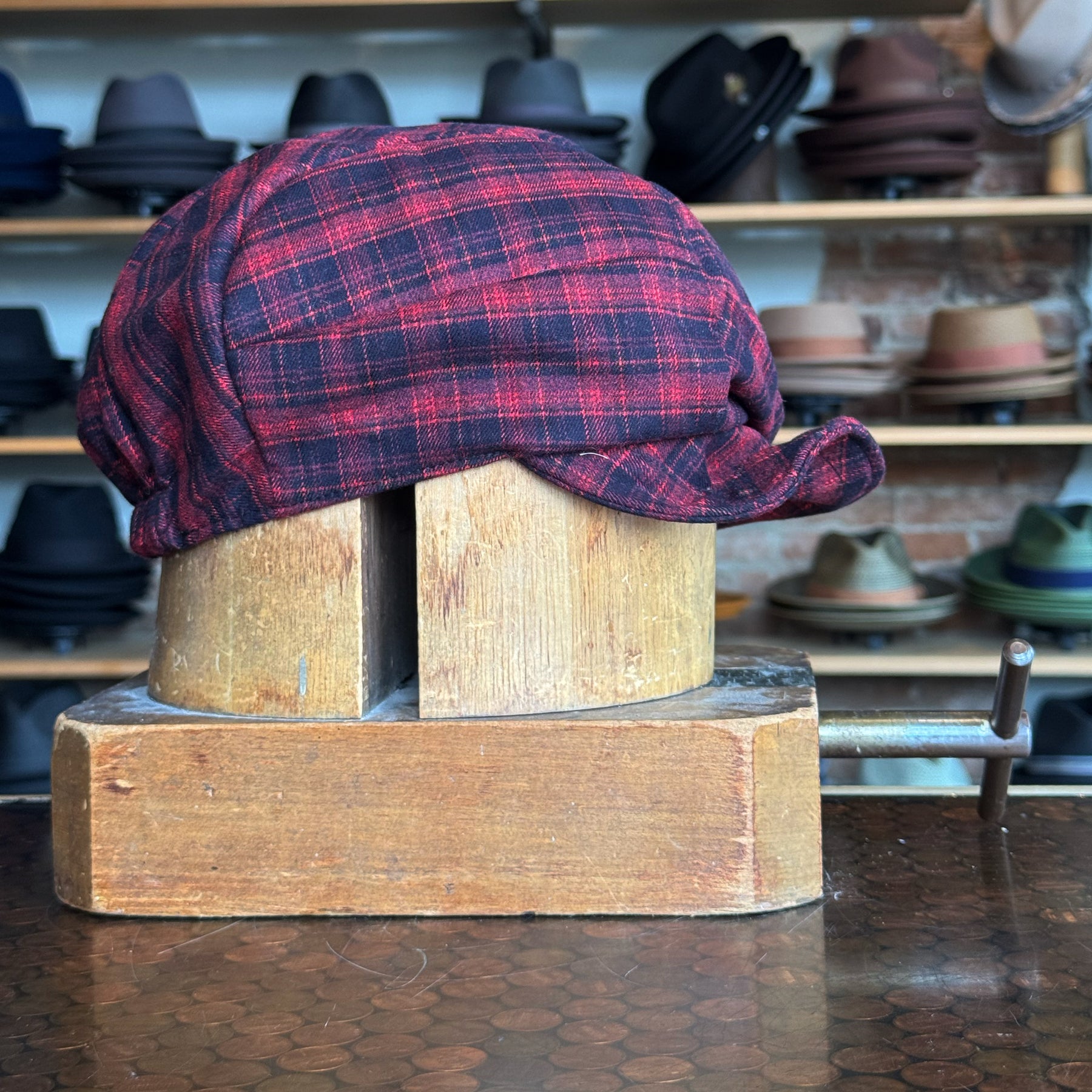Plaid Funk Master Cap in red and black plaid pattern, styled on a wooden hat block with shelves of various hats in the background, showcasing a classic newsboy cap design.