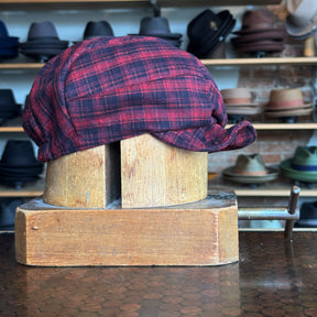 Plaid Funk Master Cap in red and black plaid pattern, styled on a wooden hat block with shelves of various hats in the background, showcasing a classic newsboy cap design.