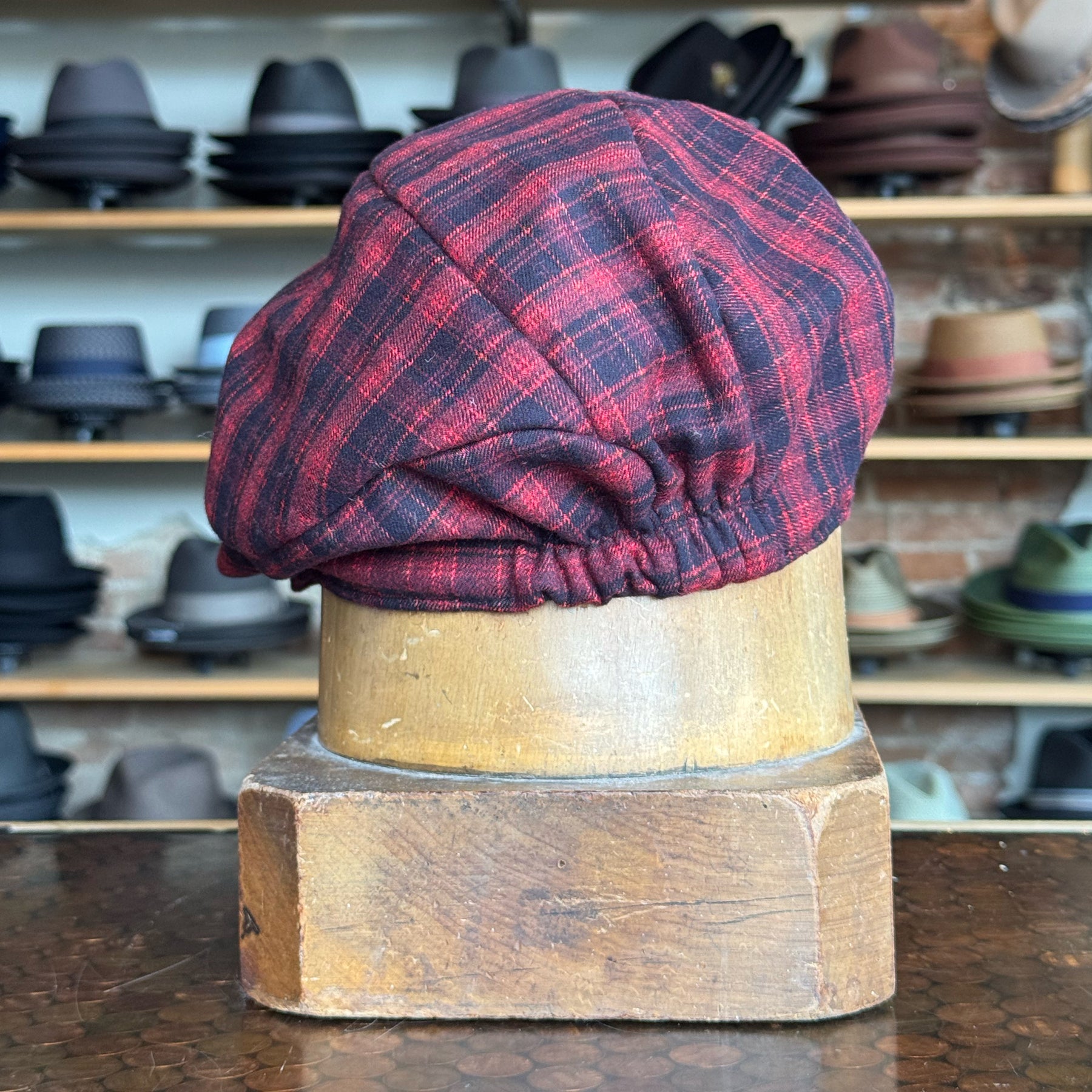 Plaid Funk Master Cap in red and black plaid pattern displayed on a wooden hat stand with various hats in the background.