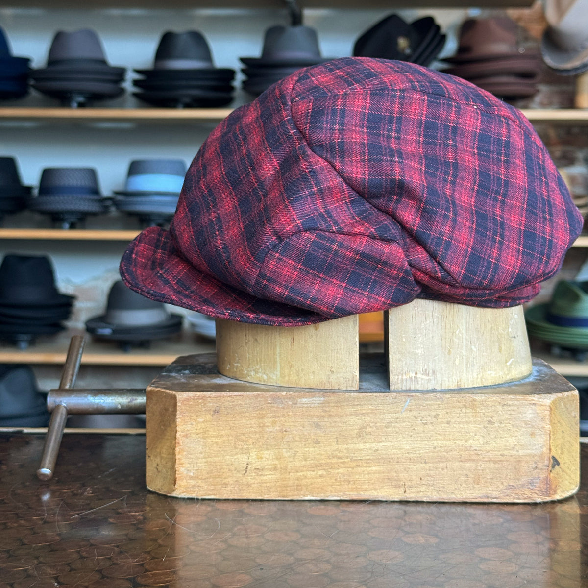 Plaid Funk Master Cap in red and black pattern displayed on a wooden hat block with a background of various hats, showcasing a stylish newsboy flat cap style.