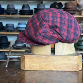 Plaid Funk Master Cap in red and black pattern displayed on a wooden hat block with a background of various hats, showcasing a stylish newsboy flat cap style.
