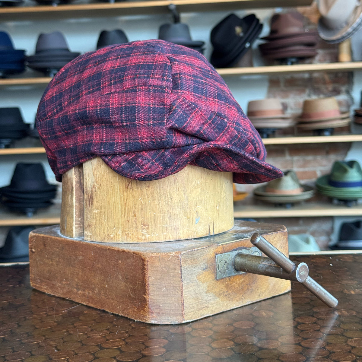 Plaid Funk Master Cap in red and black plaid pattern displayed on a wooden hat block, surrounded by various styles of hats on shelves in the background.